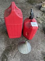 Two red plastic gas containers and one metal funnel placed on the ground, one container is the Coleman Gasoline brand.