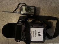 Photo showing the walkie talkie with antenna and carrying case with shoulder strap laid on floor.