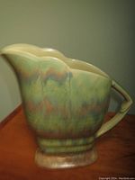 Side view of ceramic pitcher showing multi-tone green, blue, purple, and brown drip glaze, scalloped top edge and angular handle with geometric shape.