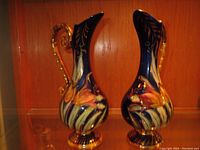 Front and side view of two tall antique Belgian porcelain vases with floral decorations and gilded handles, one with repaired handle.