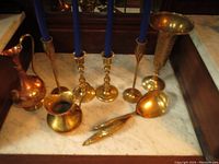 Assorted brass and copper items on marble surface including candlesticks, vases, spoons, and opener.