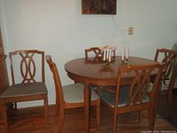 Dining table and chairs shown in a room with wood floor. Oval table with wood base and veneer top, surrounded by five chairs with wooden frames and fabric seats.