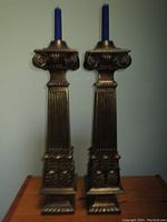 Front view of two ornate Roman column style bronze candlestick holders with dark blue taper candles inserted.