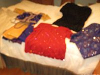 Blurry photo showing five garments spread on a bed: purple gold sari fabric, red beaded top, black beaded top, purple beaded top, and blue beaded Salwar Kameez.
