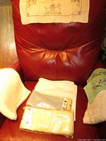Photo shows folded white cotton tablecloth, packages of cotton napkins, embroidered antimacassar draped over chair back, and folded embroidered tea towels.