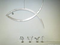 Photo showing metal fish shaped mobile hanging against wall with four small wire sculpted pin people dancer figures lined up in front on a surface.
