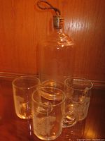 Photo shows the glass decanter with a cork stopper and attached string, alongside three clear glass mugs with etched designs.