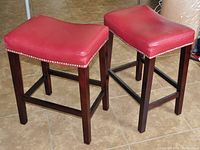 Pair of two bar stools with red leather seats and dark wood legs shown from side angle on tile floor.