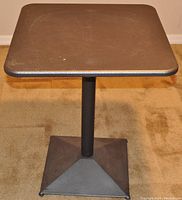 Full view of the adjustable heavy duty metal table showing square top, central cylindrical column and pyramid-shaped base, placed on beige carpet.