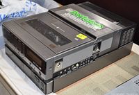 Black and gray portable VCR unit with VHS cassette slot, playback buttons, and multiple ports, laid on a flat surface with visible branding and condition sticker.