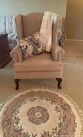 Beige upholstered armchair with floral pillow and light throw blanket on seat in room setting with visible carpet flooring. The round rug is placed in front of the chair.