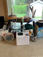 Kitchen items including bread maker, metal cooking pots, graters, glass pie dish, wooden handled utensils on a clear table in front of window.