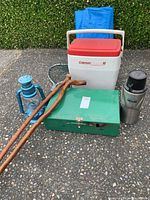 Full view of camping items arranged on pavement showing Coleman cooler, folded blue tarp, green camping stove, blue lantern, silver thermos, and two walking sticks.