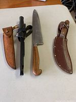 Three cutting utensils with two leather sheaths on a beige surface
