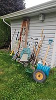 Wide view showing all garden tools arranged against a siding wall, including various shovels, spades, rakes, a snow shovel, a green garden cart with cloth cover, and a wooden ladder leaning on the wall. A bird house is mounted on the wall but explicitly not included in sale.