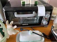 Front view of black Proctor Silex toaster oven, ceramic napkin holder with bunny, butter dish with lid, and a portion of Keurig coffee machine on the side.