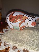 Porcelain pig figurine with orange, brown, and green painted design, shown on a crocheted cloth.