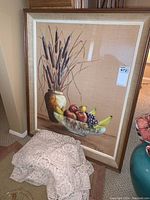 Framed artwork showing fruit bowl and vase with cattails beside beige lace tablecloth partly draped