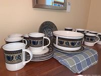 Wide view of Mikasa Arabella Intaglio dinnerware set including several coffee mugs, saucers, and a large serving bowl on a wooden table with checkered cloth.