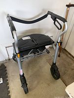 Front and side view of silver walker with built-in black padded seat and foam grip handles, showing front wheels and foldable structure.