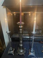 Three metal candle sticks arranged in front of a fireplace, each holding a lit candle. Tallest candle stick is centered and flanked by two shorter candle sticks. Photo shows size and condition of candle sticks.