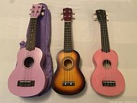 Three ukuleles on a flat surface, showing colors and size differences; one pink with carry bag, one sunburst, one pink
