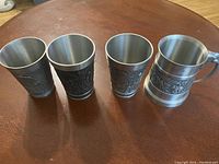 Four pewter mugs with varied detailed relief decorations in very good condition shown side by side on a wooden table.