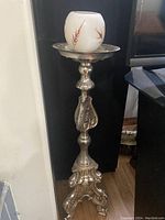 Full view of the tall metal candle stand featuring a decorative top tray holding a white candle with leaf patterns.