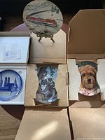 Set of four collectible plates in original boxes shown from the front with artwork visible, including a red train, dog, koala, and blue cityscape.