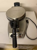 Front view showing the Waring logo on the waffle maker's round stainless steel lid and its black handle, cord visible to the right.