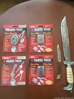 Photo showing four new Swiss Tech multi-tools in original packaging alongside two used knives with distinct handles and blades.