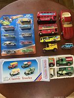 Overview image showing 8 Hot Wheels cars in original packaging, 4 classic trucks in box, 2 London-themed double-decker buses, a yellow vintage truck, a red convertible car, and a small blue car.