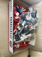 Box filled with assorted LEGO bricks and pieces, including some in plastic bags, showing variety of colors and types of bricks.