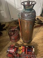 Three firefighter-related items displayed on wood floor: copper fire extinguisher, red metal gas can, and vintage red metal fire truck.