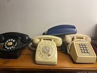 Lot of five vintage telephones including rotary dial and push-button models on wooden surface against white wall.