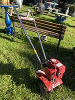 Red Mantis rototiller positioned on green grass, showing overall appearance with metal handles, red engine housing and tines for soil cultivation.