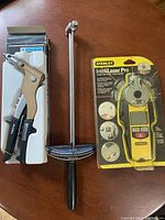 Three tools on a wooden surface: Stanley Intelli Laser Pro in original packaging, a Mastercraft heavy duty hand riveter also in box, and an unboxed torque wrench with adjustable scale and black handle.