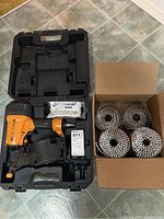 Stanley Bostitch brad nailer in open carrying case alongside cardboard box of coil nails.