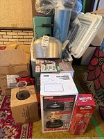 Photo showing cooler, kettle, boxed Coleman lantern, and boxed Garrison lantern along with other camping items.