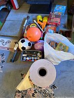 Wide view of the lot showing balls, puzzles, books, toy guns, a white bag of puzzle boards, and other miscellaneous items on a carpeted floor.
