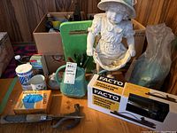 Image showing garden figurine, FACTO oscillating sprinkler box, green watering can, and assorted garden tools and items on wooden floor.