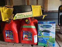 Two red fuel cans with 5.3 gallon capacity, Hydro Breeze 3-in-1 spreader in box, and a yellow box labeled Vinylex Lawn Edging on a wooden shelf.