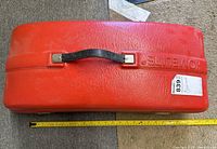 Closed red plastic Homelite branded hard carrying case containing the two chainsaws, shown with tape measure for size reference.