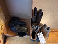Photo of multiple pairs of men's boots in a cardboard box, including black leather CSA certified boots and other tan and black boots.