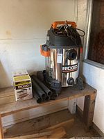 Front view showing Ridgid shop vac stainless steel body, tubes, and replacement filter box on wooden table.