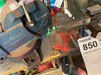 Two metal bench vices on a workbench. One blue and one red, both showing signs of rust and wear, with mounting bases visible. Blue vice attached to bench.