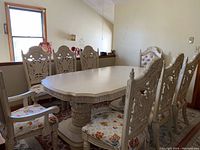Full view of the dining table and some chairs showing detailed wood carving and white finish.
