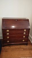 Full front view of the antique wooden secretary desk closed, showing five rows of drawers and the slant-top drop front door with brass handles.