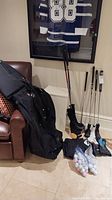 Photo showing large black Club Glove XXL travel golf bag leaning on chair with golf clubs, head covers, and bags of golf balls on floor