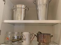 Two shelves showing three metal ice buckets, ceramic and metal steins, three glass jars, and a copper-colored bowl.